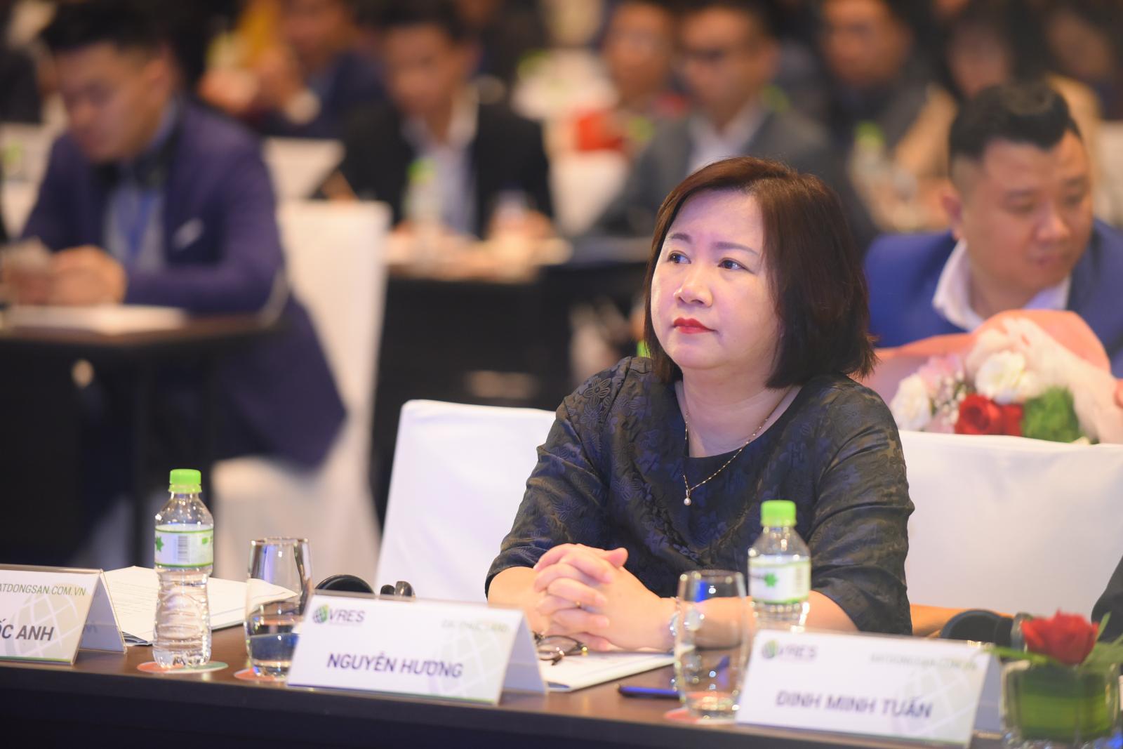 Chân dung một phụ nữ trung tuổi mặc váy đen, tóc ngắn, ngồi giữa hội thảo.