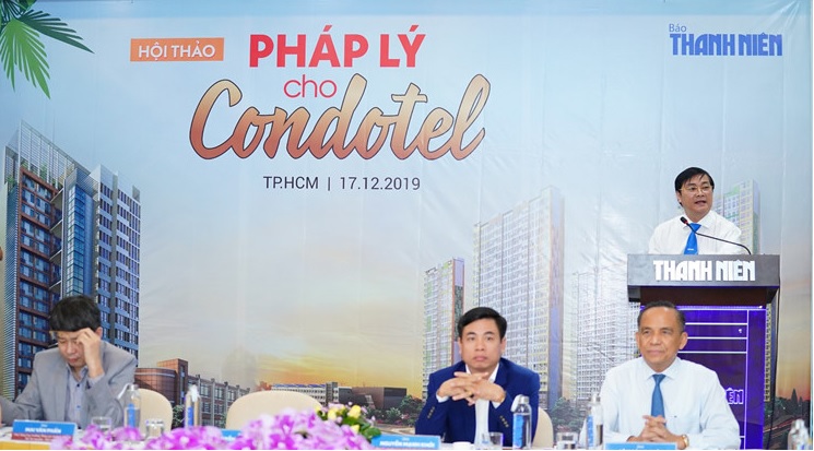 Ba người đàn ông luống tuổi ngồi trên bàn gần phông nền sân khấu, một người đứng trên bục phát biểu tại hội thảo về condotel.